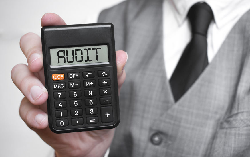 Person in a gray vest and tie holding a calculator displaying "AUDIT" on the screen.